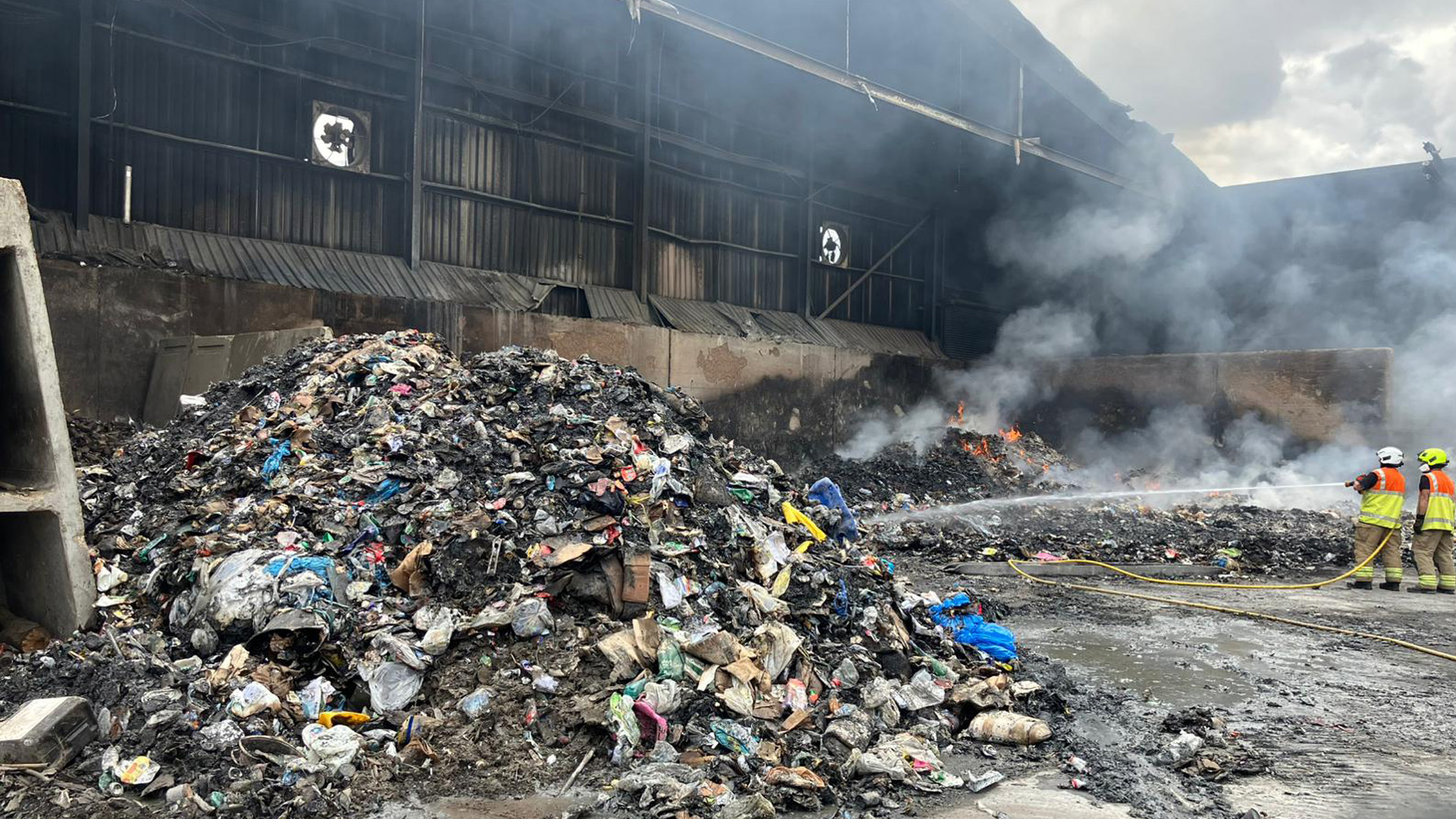 Two firefighters point hose reels at a large pile of smouldering rubbish at the Elstow Waste Transfer Station fire. 