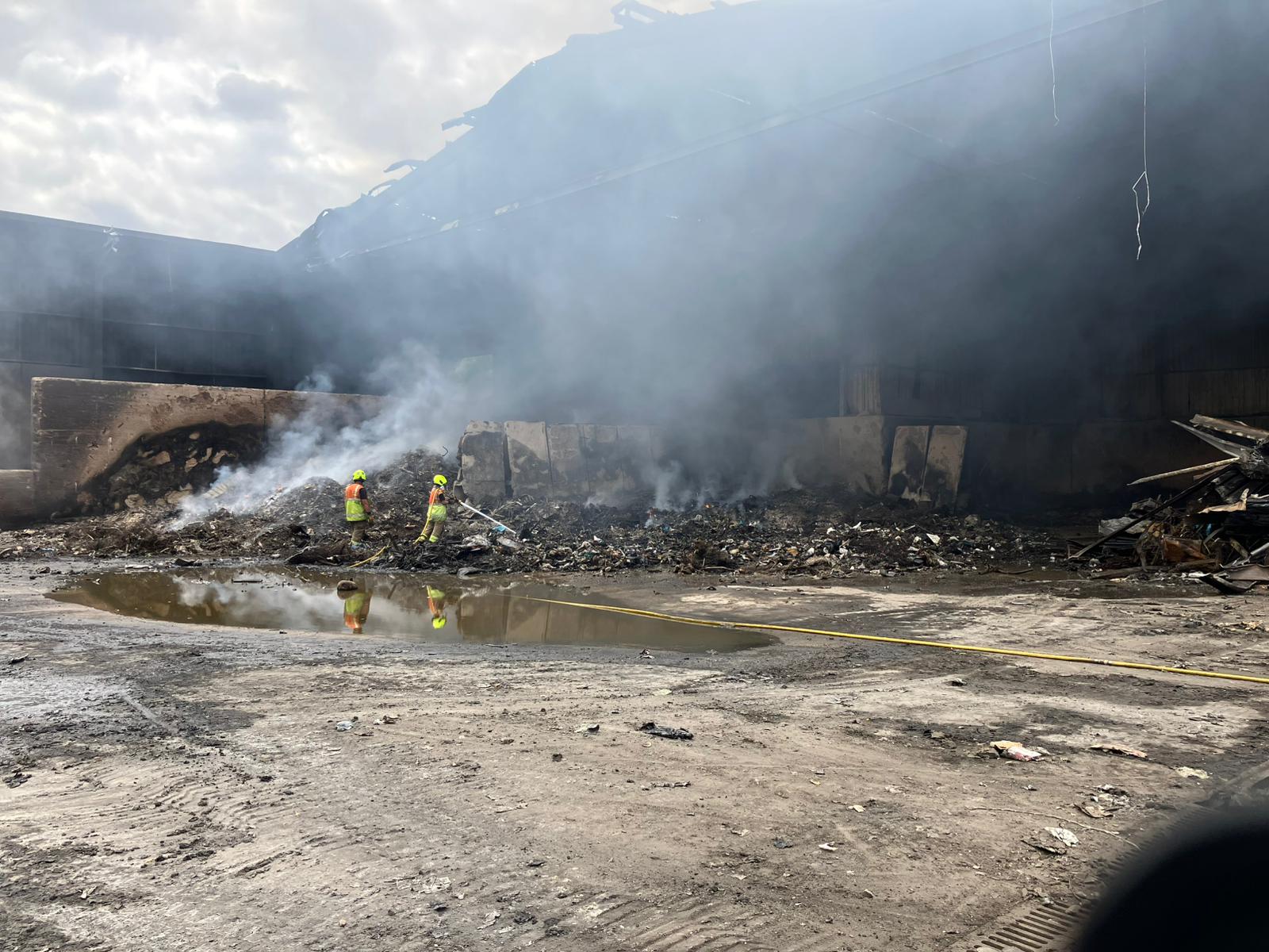 Two firefighters stand in front of a smouldering pile of rubbish, one pointing a hose reel towards the rubbish.
