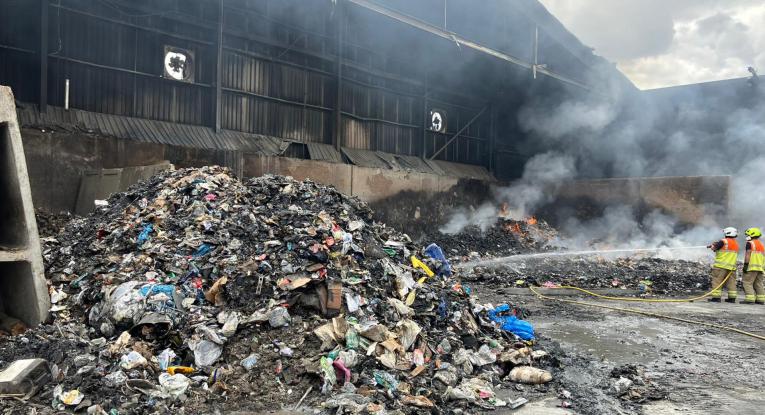 Two firefighters point hose reels at a large pile of smouldering rubbish at the Elstow Waste Transfer Station fire.