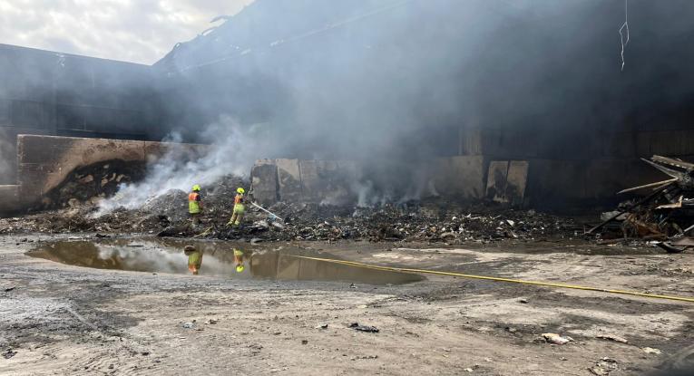 Two firefighters stand in front of a smouldering pile of rubbish, one pointing a hose reel towards the rubbish.